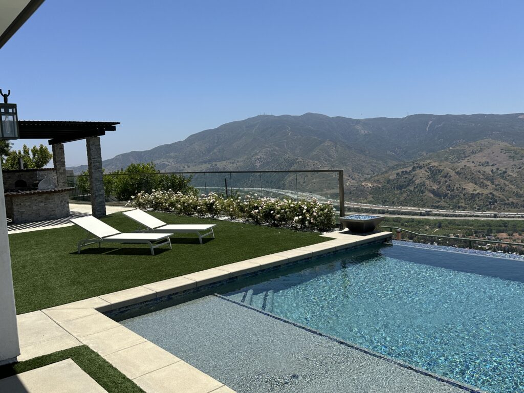 Photo of a modern backyard in Yorba Linda, CA.  The photo shows an infinity pool, grassy area, and chaise lounges.  There is an outdoor glass railing system bordering the edge of the backyard.