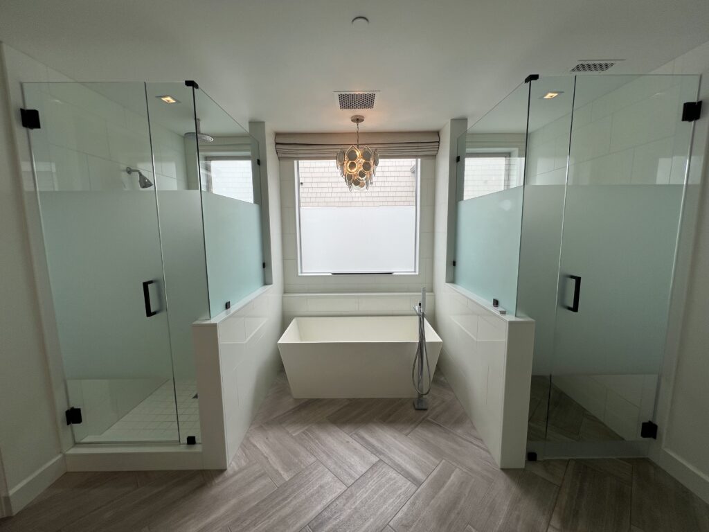 A photo of a bathroom with two showers and a bath tub.  Frosted Glass Shower Doors are used to create additional privacy for both showers. A frosted glass window is also shown in the background to create additional privacy.